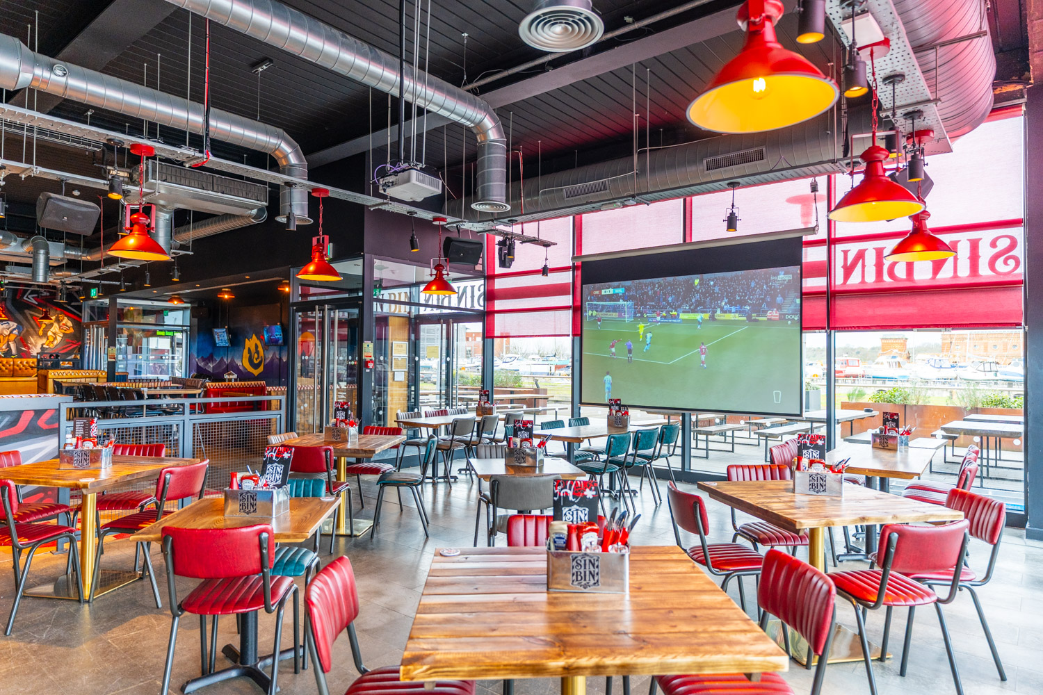 A seating area is for eating at the Sin Bin in Chatham. The tables are wooden and the seats are red, as is the lights above. The projector at the far end of the room is playing the football.
